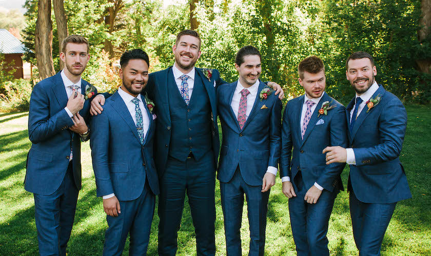 Group of men in blue suits and ties standing outdoors with greenery in the background