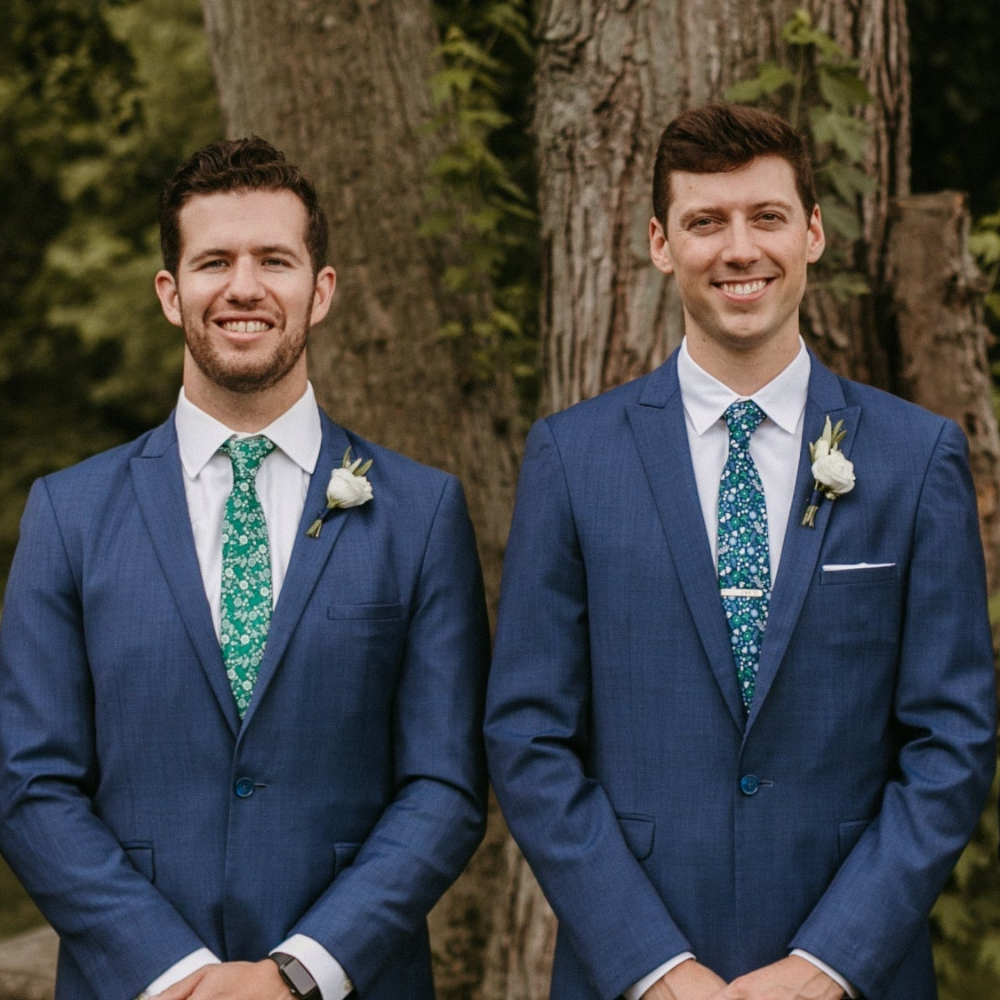 Two men in blue suits with green ties standing outdoors against a tree background